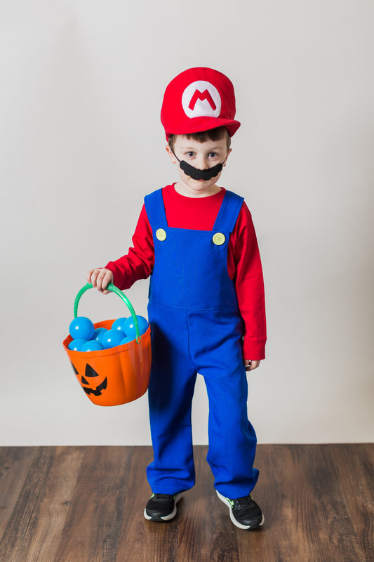 Young boy in handmade Mario Bros costume — red cap with white sewn-on M logo, red long sleeve t-shirt, blue overalls with plastic yellow buttons, and classic black mustache, holding a pumpkin candy bucket