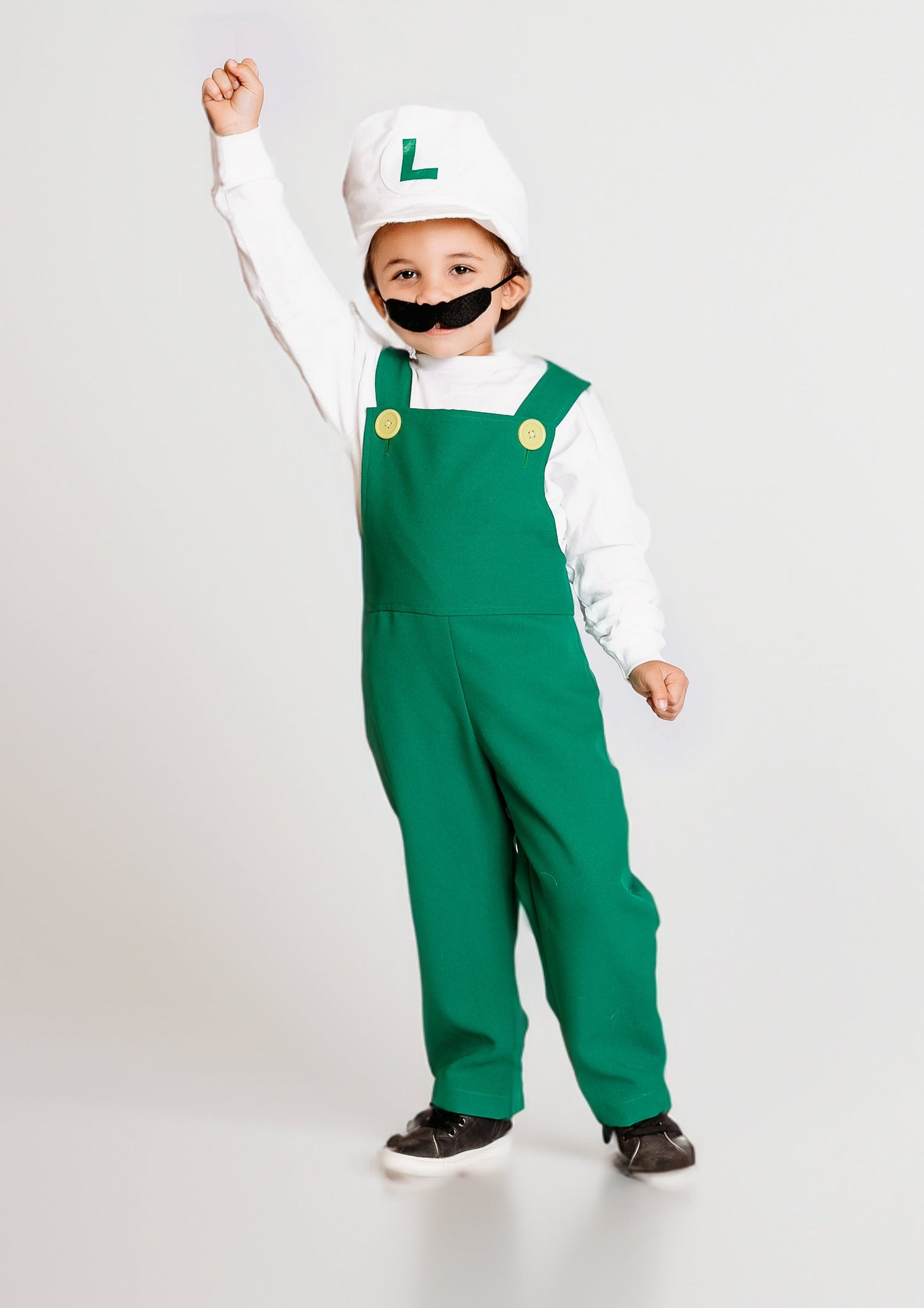 Young child celebrating with fist raised in handmade Fire Luigi Bros costume — white cap with green L logo, white long sleeve t-shirt, green overalls with yellow buttons, and black mustache