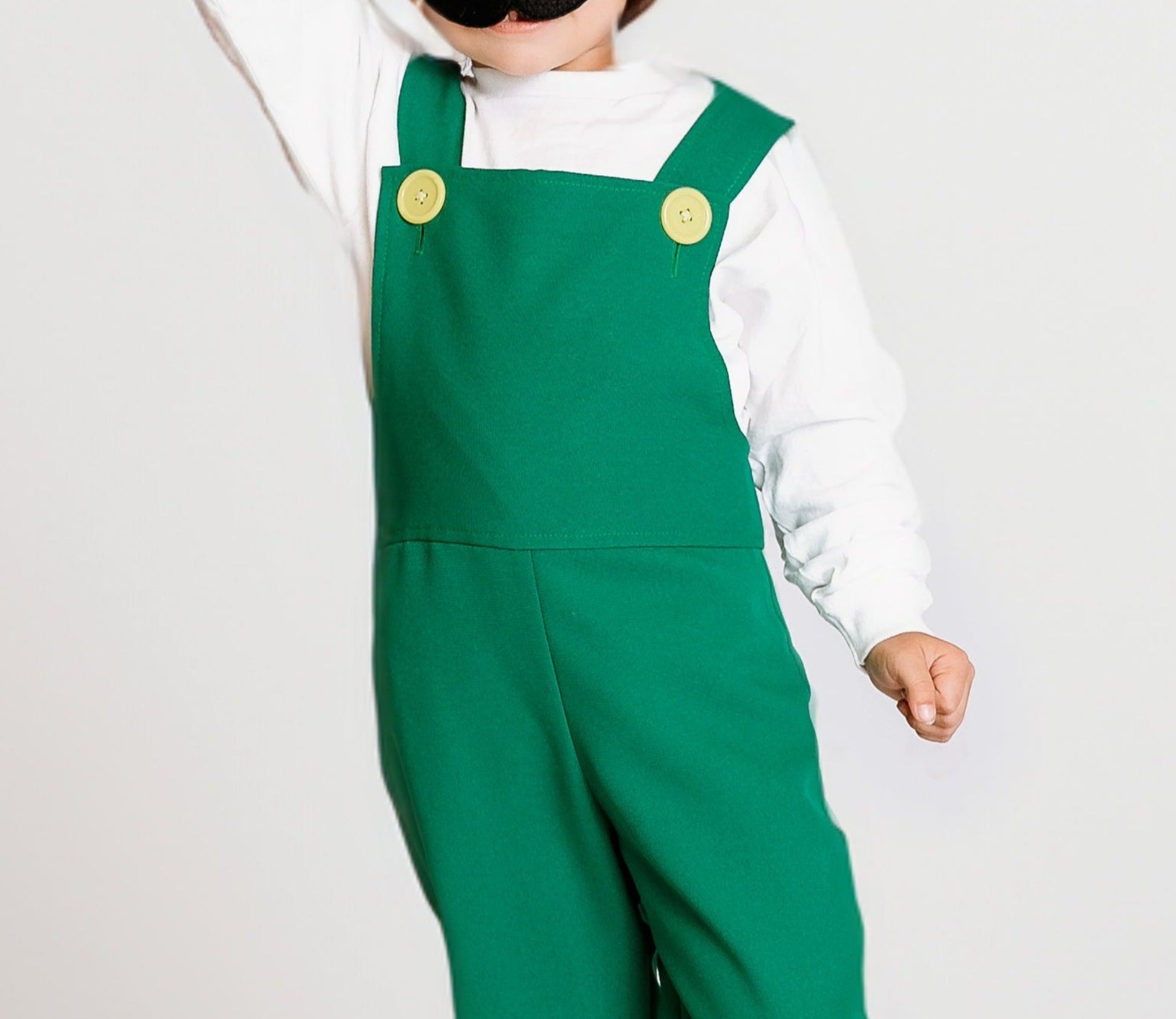 Young child celebrating with fist raised in handmade Fire Luigi Bros costume — white cap with green L logo, white long sleeve t-shirt, green overalls with yellow buttons, and black mustache