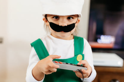 Child in handmade Fire Luigi Bros costume holding a video game controller — white cap with L logo, white long sleeve t-shirt, green overalls with yellow button, and black mustache