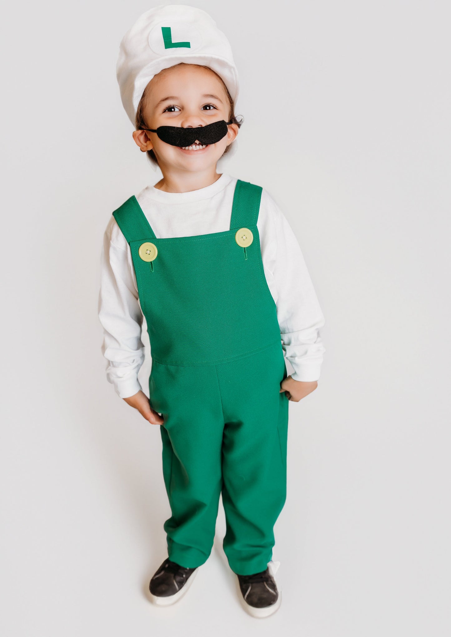 Smiling toddler in handmade Fire Luigi Bros costume — white cap with green L logo, white long sleeve t-shirt, green overalls with yellow buttons, and black mustache