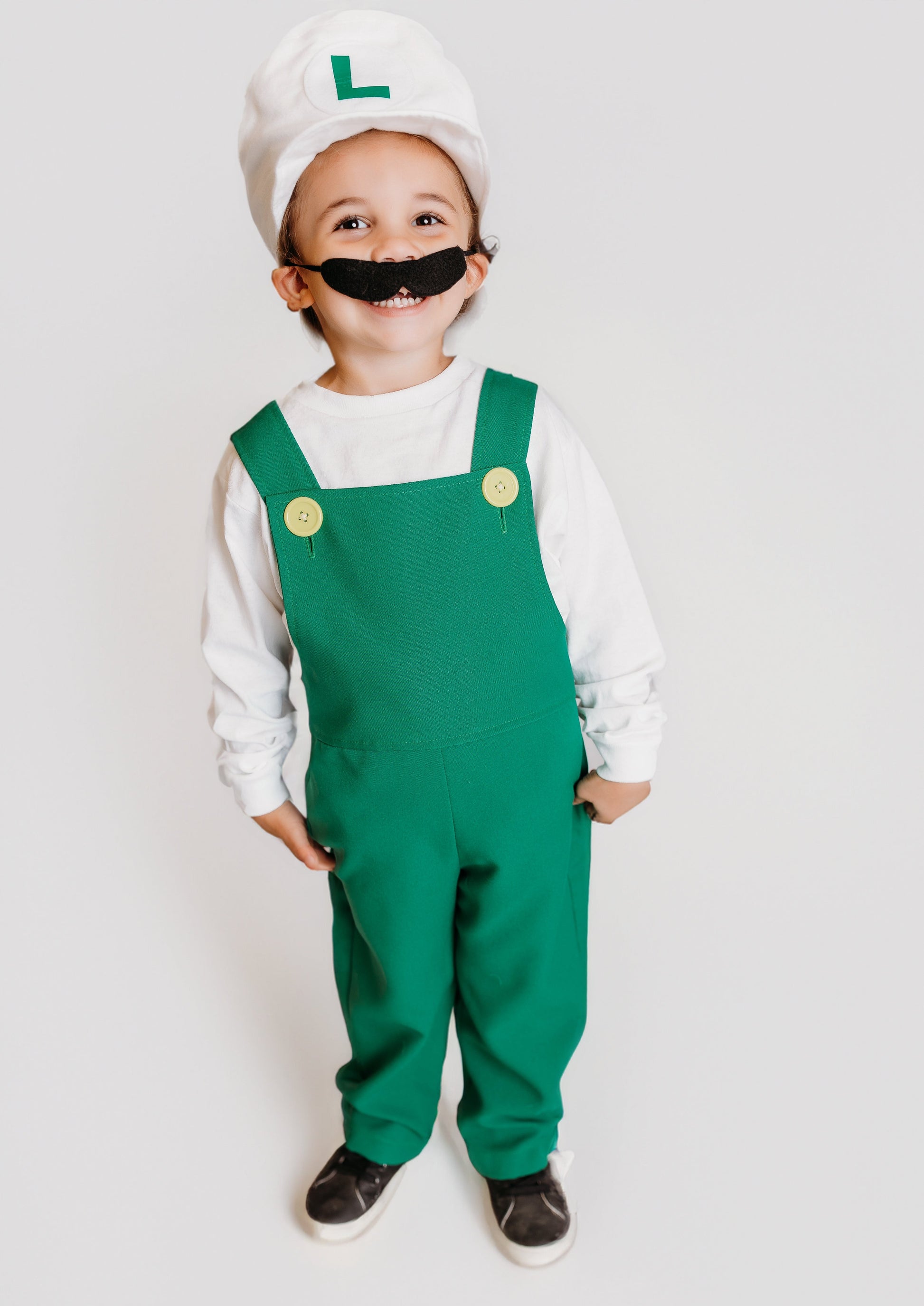 Smiling toddler in handmade Fire Luigi Bros costume — white cap with green L logo, white long sleeve t-shirt, green overalls with yellow buttons, and black mustache
