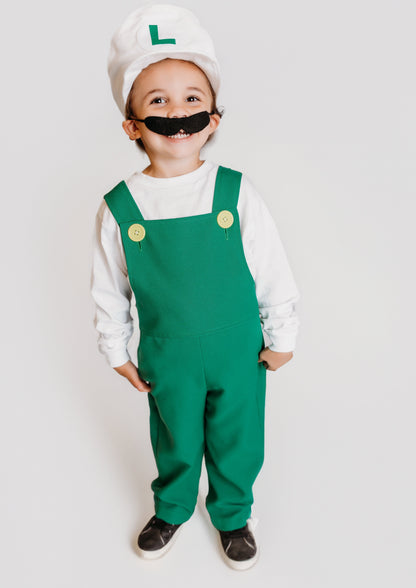 Smiling toddler in handmade Fire Luigi Bros costume — white cap with green L logo, white long sleeve t-shirt, green overalls with yellow buttons, and black mustache
