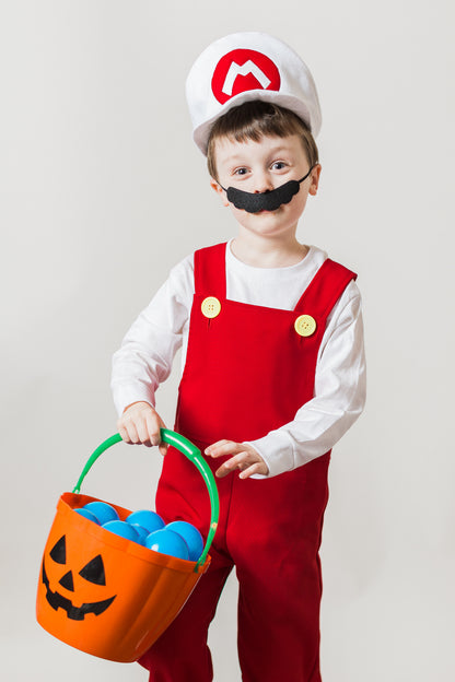Young boy in handmade Fire Mario Bros costume — white cap with red M logo, white long sleeve t-shirt, red overalls with yellow buttons, and black mustache, holding a pumpkin candy bucket