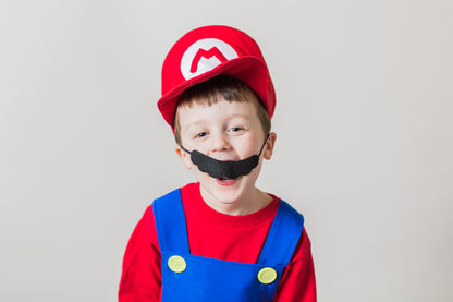 A child wearing a Mario Bros costume, featuring a red hat and blue overalls, with a black mustache accessory designed to resemble Mario's mustache.