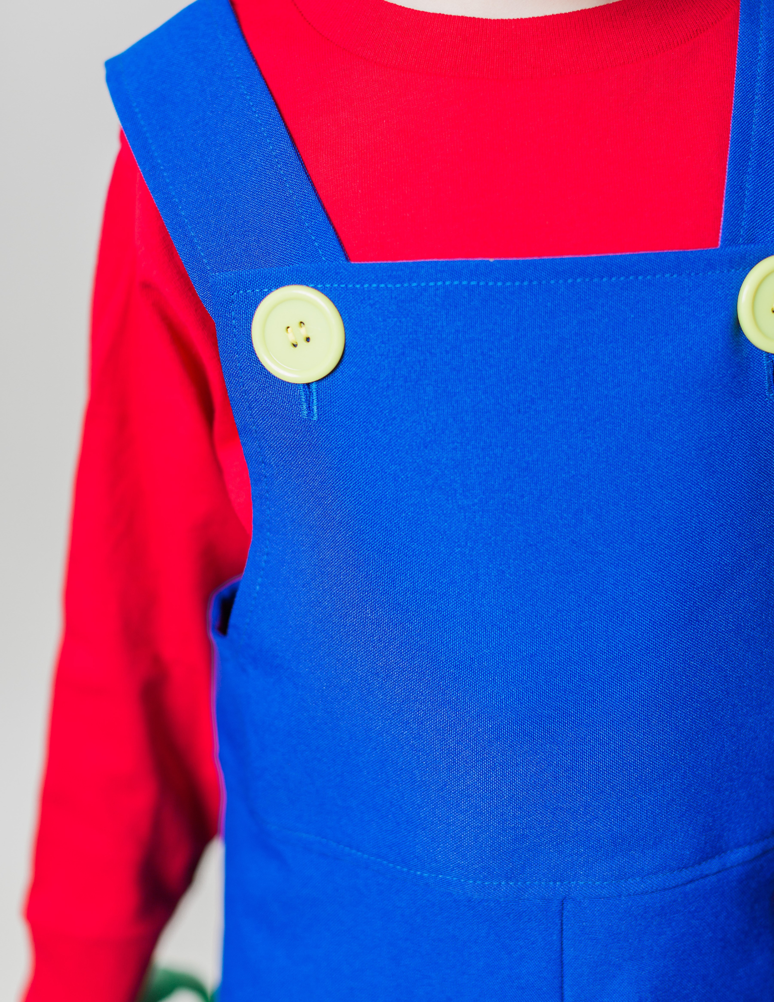 Close-up detail of handmade Mario Bros costume — blue polyester overalls with plastic yellow buttons over a red long sleeve t-shirt