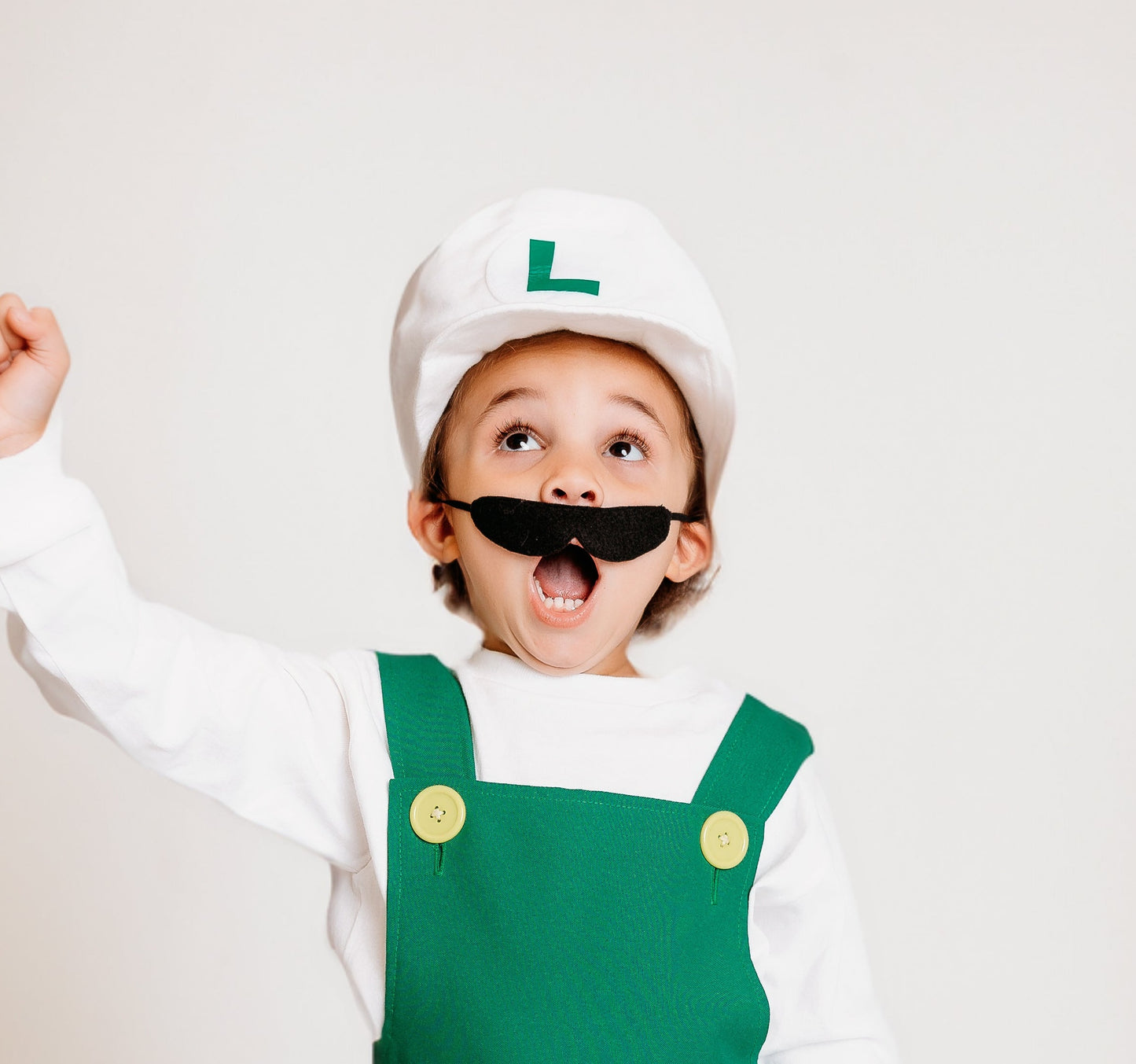 Excited child in handmade Fire Luigi Bros costume — white cap with green L logo, white long sleeve t-shirt, green overalls with yellow buttons, and black mustache, with fist raised in celebration