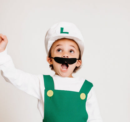 Excited child in handmade Fire Luigi Bros costume — white cap with green L logo, white long sleeve t-shirt, green overalls with yellow buttons, and black mustache, with fist raised in celebration