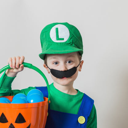 Smiling boy in handmade Luigi Bros costume close-up — green cap with white L logo, red long sleeve t-shirt, blue overalls with yellow buttons, and classic black mustache holding  Halloween bucket with blue small plastic balls